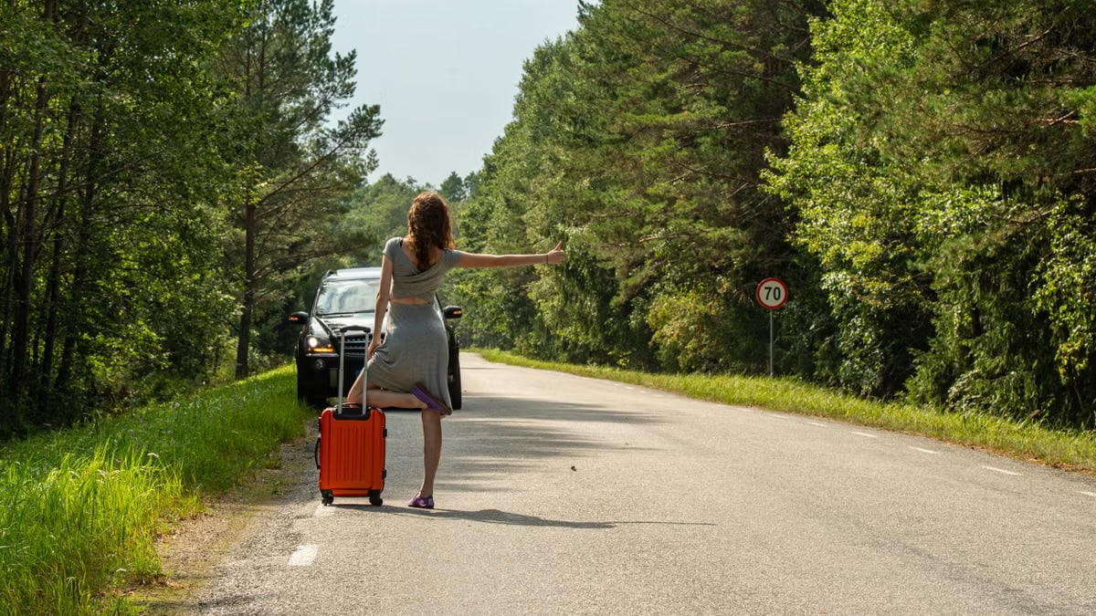 A woman hitchhiking with an orange suitcase on a rural road in Estonia during summer.